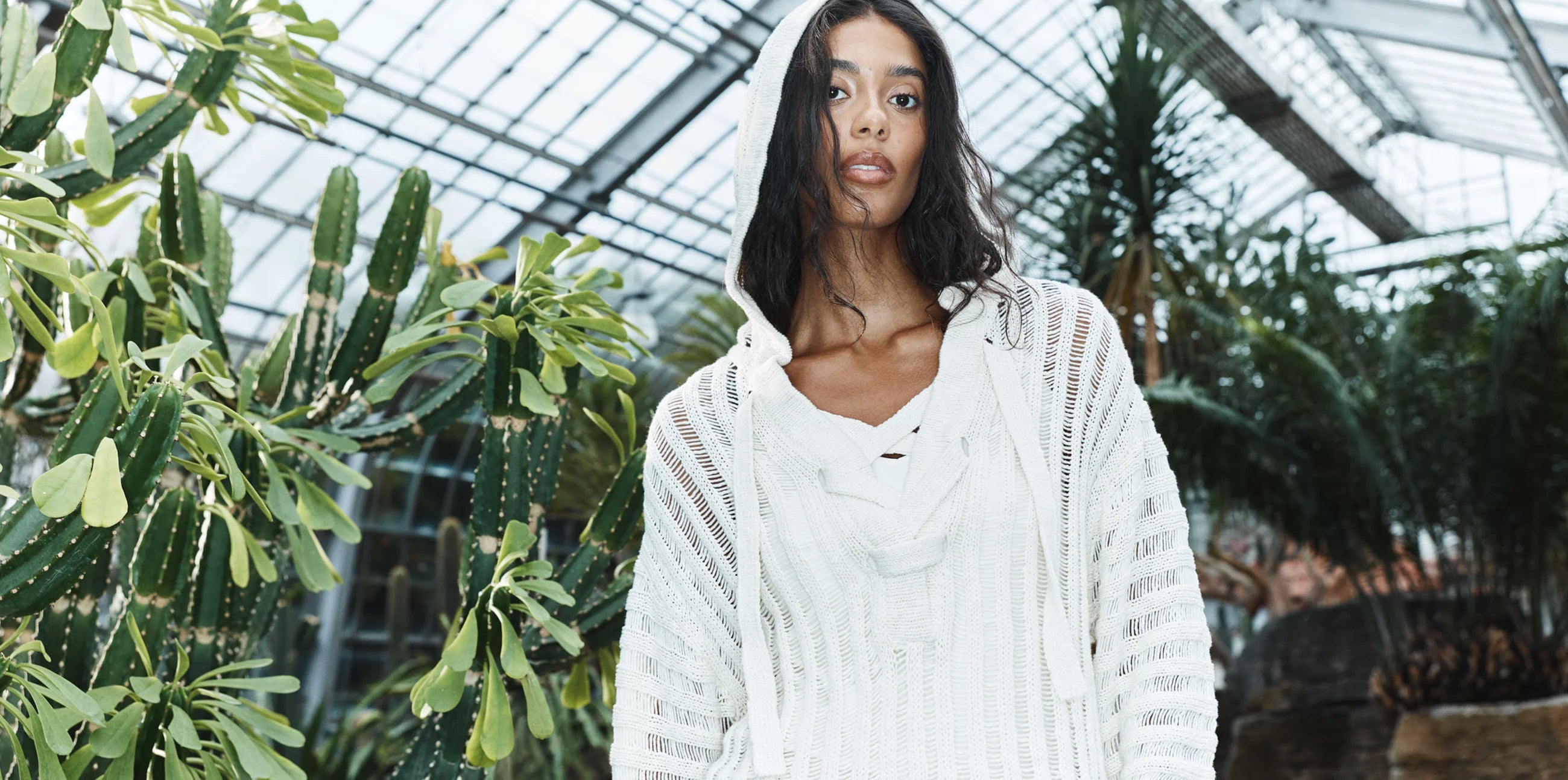 Femme portant un chandail à capuchon en tricot ajouré blanc JOELLE, dans une serre remplie de cactus et de plantes tropicales